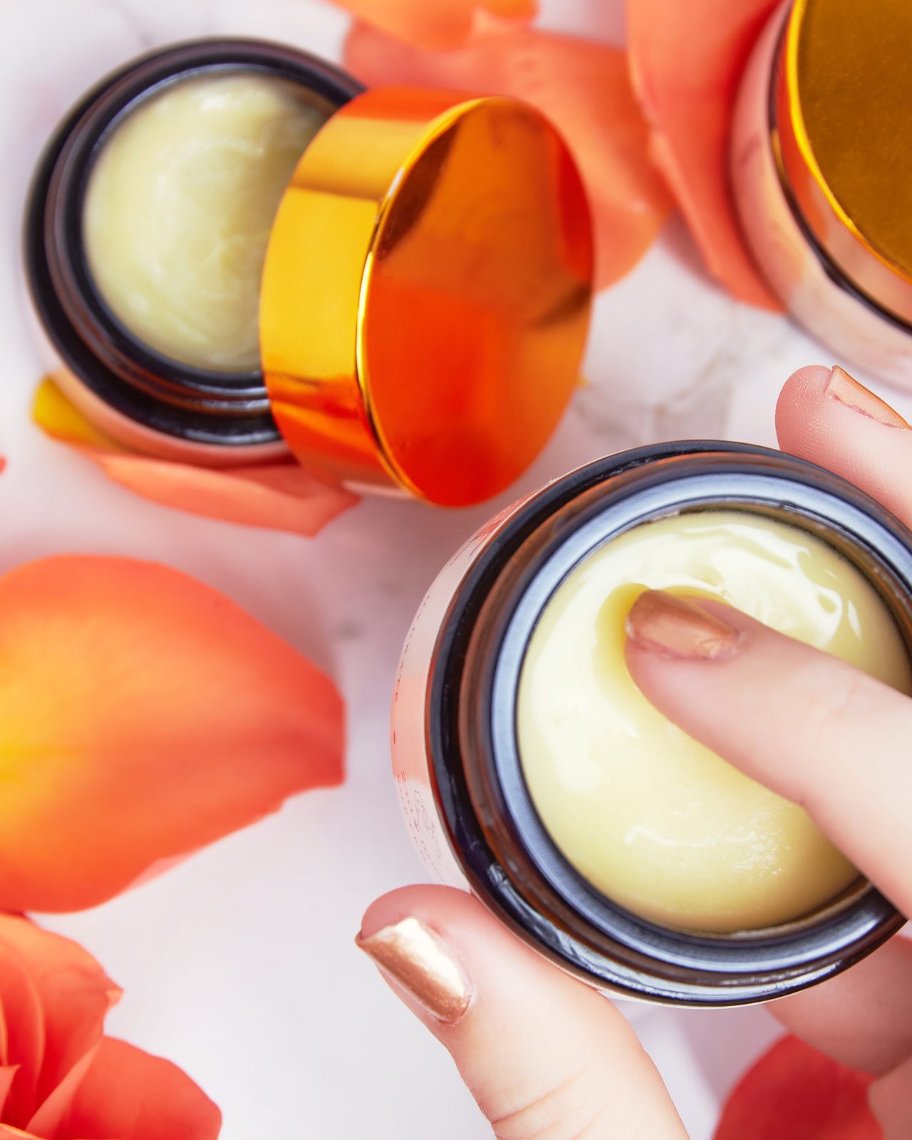 A finger dipping into an open jar of Pussy Butter™ among coral rose petals